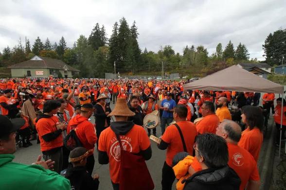 A large group of people are seen, almost all wearing orange shirts, in a large parking lot. A circle of young and old men drum and sing. Visible in the picture are the Wawmeesh Ken Watts elected chief of Tseshaht, (second from left with cedar headband), Brandy Lauder elected chief of Hupacasath (bottom right closest to camera, black jacket), Gord Johns MP for Courtenay Alberni (balding grey hair beside Lauder), and Mayor Sharie Minions of Port Alberni (dark straight hair beside Gord Johns).