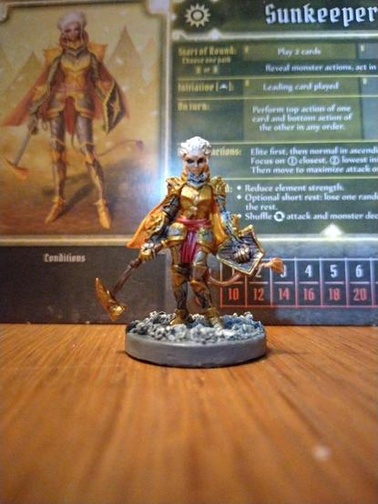 Mini of the sunkeeper from Gloomhaven painted with the intention of matching the character art (displayed in the background)