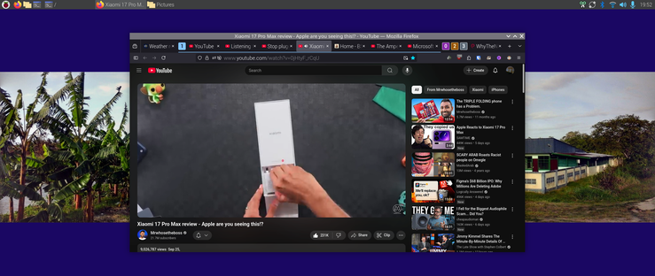 The image shows a computer screen with a YouTube video playing in the center. 
The 100W charger is promintently displayed here
The video is titled "Xiaomi 17 Pro Max review - Apple are you seeing this?" and features a close-up of a hand holding a black smartphone displaying a smaller video on its screen. The video has 21.1M views and was uploaded on September 23. The YouTube interface includes a search bar, a sidebar with related videos, and a play button. The browser tabs at the top include "Weather," "YouTube," "Listening," "Stop," and "Xam...". On the left side of the screen, there is an image of banana trees, and on the right, there is a picture of a house with a blue roof and white walls. The background of the screen is a dark blue color.