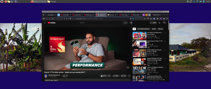 The image shows a computer screen with a YouTube video playing in the center. 
Snapdragon 8 is highlighted here
The video is titled "Xiaomi 17 Pro Max review - Apple are you seeing this?" and features a person opening a white box. The video has 9,526,776 views and was uploaded on September 25. The YouTube interface includes a search bar, a sidebar with related videos, and a channel name "MehtheosTheSethos." The browser tabs at the top include "Xiaomi 17 Pro Max review - Apple are you seeing this?" and "Weather." On the left side of the screen, there is an image of banana trees, and on the right side, there is an image of a house with a blue roof and a white fence. The time displayed on the screen is 19:19.