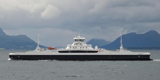 A photo of a large black and white ferry crossing a body of water with mountains in the distance. The sky is filled with clouds.