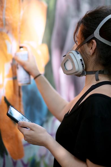 A streetart female artist painting with spray can and mask, with her phone in her left hand. She is focused on the mural she is painting. The wall and photo tone is orange and warm.