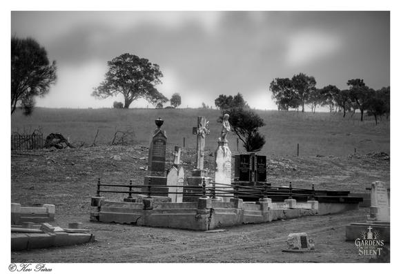 A wide, atmospheric black and white photograph of an old, rural cemetery in Tungkillo, South Australia.

In the foreground, a prominent plot is enclosed by low concrete and wrought iron railings, featuring several historic headstones.

One tall, intricately carved monument stands centrally, and another to its right is topped with a small angelic figure. 

The ground is dry earth and patchy scrub. In the mid-ground, a large, gnarled tree dominates the horizon line, set against a rolling, barren field.

The background features a dense line of darker trees and a sky heavily blanketed in soft, bright mist or fog, creating a deep sense of stillness and isolation.