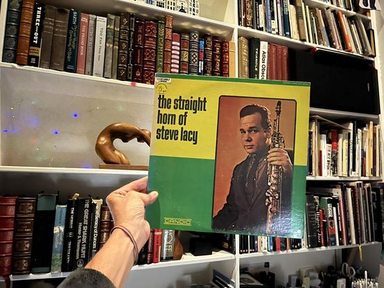 This photo shows someone [me again] holding a vintage jazz album titled "The Straight Horn of Steve Lacy" on the Candid label. The album cover has a distinctive color-blocked design with a bright yellow section at the top and green sections at the bottom. On the right side of the cover is a photo of Steve Lacy in a dark suit holding a soprano saxophone against an orange background. There's a small circular label in the upper left indicating "GREAT EARLY CLASSICS."

The setting appears to be a well-stocked home library, with extensive bookshelves visible in the background filled with leather-bound volumes and various hardcover books. The shelves are white and well-organized, containing books of different colors including burgundy, brown, black, and other tones.

On one of the shelves behind the album, there's an interesting wooden sculptural object that appears to be curved or abstract in form. There are also some small colored lights (appearing as blue and multi-colored dots) creating an ambient atmosphere in the background. A bookshelf speaker is also visible mounted among the bookshelves.

The person holding the album [👋🏽] is [still] wearing a [friendship] bracelet on their wrist [where else?], similar to the previous photo. [ALT mostly by Claude]