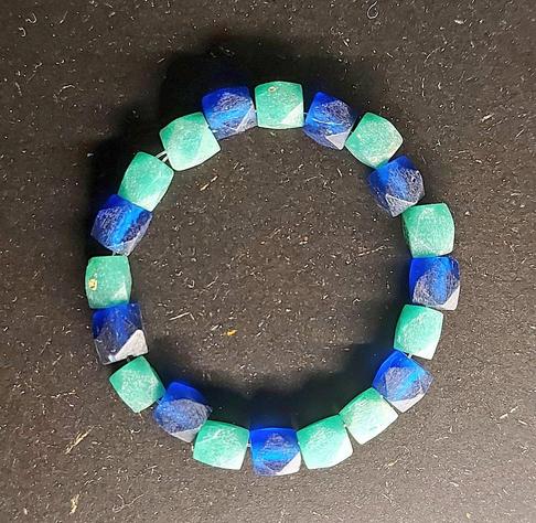 A beaded bracelet made of alternating cube-shaped stones in turquoise green and deep blue, arranged in a circle on a dark textured surface.