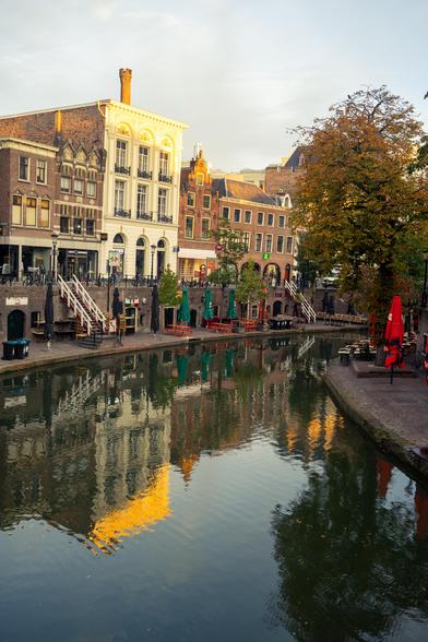 The image depicts a picturesque canal scene in what appears to be a European city. The canal, calm and reflective, runs through the center of the image, mirroring the surrounding buildings and trees. On the left side of the canal, there are several multi-story buildings with distinct architectural styles. The building closest to the viewer is a white structure with ornate details, featuring large windows and a chimney on the roof. Adjacent to it are other buildings with brick facades and varying window designs.

Along the canal, there are outdoor seating areas with tables and chairs, some shaded by green umbrellas. A few people can be seen sitting and walking around, suggesting a lively atmosphere. On the right side of the canal, there is a tree with autumn-colored leaves, adding a touch of nature to the urban setting. The sky above is clear with a hint of clouds, and the overall lighting suggests it might be early morning. The scene is tranquil and inviting, capturing the charm of a canal-side setting.