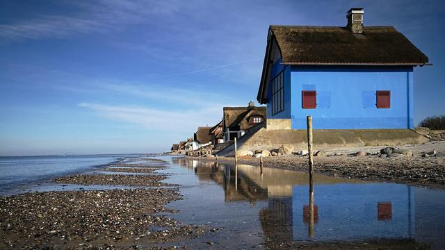 das bild im format 16:9 zeigt eine reihe von strandhäusern mit reetdächern, die entlang der küstenlinie stehen. im vordergrund befindet sich ein strandbereich mit wasser, das sich in kleinen lacheln sammelt und die häuser und den klaren blauen himmel darüber reflektiert. das erste haus im blick ist auffällig blau mit roten fensterläden und einem dunklen reetdach. die anderen häuser setzen sich in einer linie fort und scheinen ähnliche traditionelle reetdächer zu haben. das wasser des meeres erstreckt sich bis zum horizont, wo man verschwommen eine art brücke oder konstruktion erkennen kann. es ist ein sonniger tag, und das licht spiegelt sich im wasser und auf den fassaden der häuser. der strand besteht aus sand und kleinen steinen.