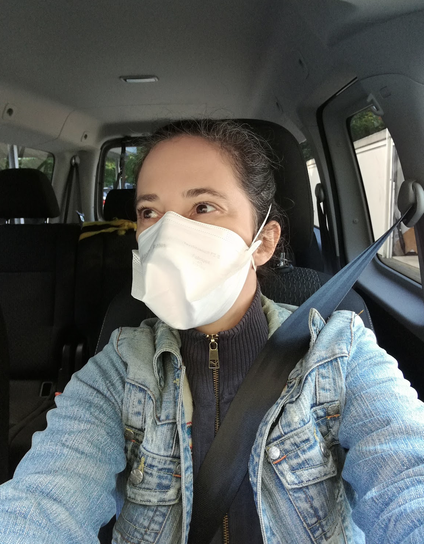 selfie of a white woman in a car. Her brown hair is pulled back. She is wearing a white duckbill respirator, a light blue denim jacket and a dark blue zippered turtleneck cardigan. She is checking the rearview mirror.
