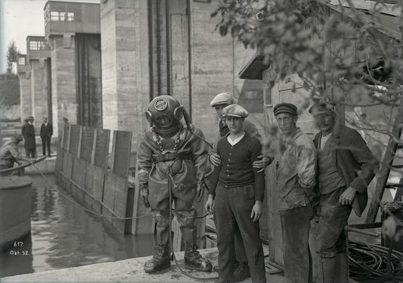 Schwarz-weiß Aufnahme: Ein Taucher und daneben vier Männer mit Mützen auf dem Kopf stehenneben einer Schleuse. Im unteren linken Eck der Vermerk Okt. 32.