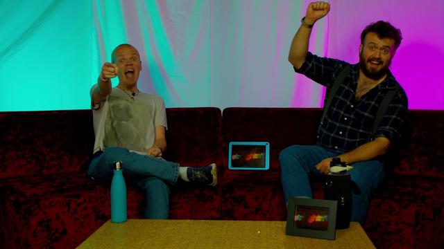 Two excited individuals are sitting on a red velvet couch, one pointing enthusiastically while the other raises a hand in celebration. A colorful backdrop and a small table with a water bottle and a framed item are visible. An electronic device displaying the "Straightn’t" logo is between them