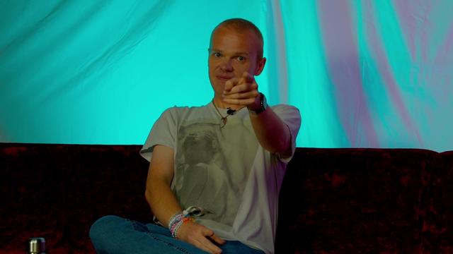 A person with short hair sitting on a couch, wearing a graphic t-shirt with Taylor Swift on it and denim jeans. They are pointing playfully at the camera with a smile. The background features a blue-green gradient.