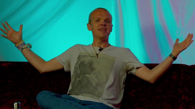 A person seated on a red couch, smiling and gesturing with both arms raised. They are wearing a light-colored t-shirt with an image of Taylor Swift on it and blue jeans. The background features a turquoise backdrop, creating a vibrant atmosphere.