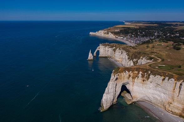 Aerial view of white chalk cliffs along a coastline with a blue ocean, featuring distinctive rock formations and a small beach area. In the background, a town is visible.