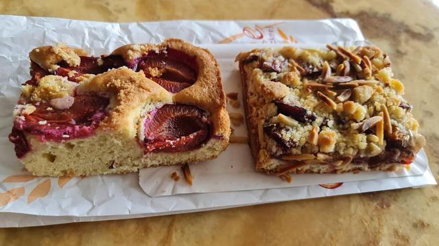 Zwei verschiedene Stück zwetschgenkuchen nebeneinander auf dem pappstück und papiertüte auf marmorierter tischplatte. Rechteckige blechkuchen, beim einen halbe Zwetschgen obenauf, beim anderen noch Streusel und Krokant, mandelsplitter obendrüber.