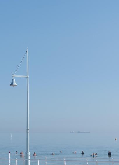Auto-generated description: People are swimming in calm waters under a clear blue sky near a street lamp, with a ship visible in the distance.