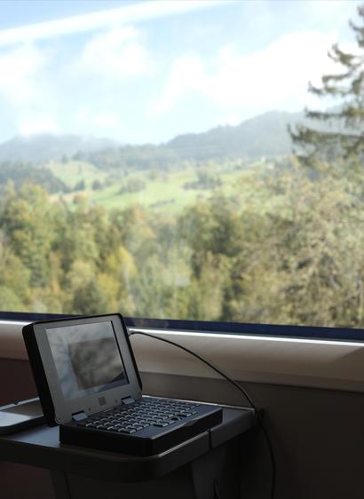 A pocket reform computer on a small table in a train. Through the window a blurry landscape of hills covered by forest and fields. Some trees are starting to turn yellow.