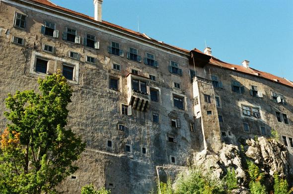 A photograph of part of the castle in Český Krumlov, Czech Republic. The picture shows a towering, weathered stone wall filled with dozens of windows in various sizes, many with wooden shutters, some of them open. A small wooden balcony juts out high above the town, giving the impression of medieval watch posts. The wall looks aged and rough, carrying centuries of history in its surface. Below, rocky cliffs merge with the base of the castle, while green trees and bushes climb upward, softening the heavy stone. Above it all, the roof with red tiles and chimneys lines the top edge of the structure. The sky is a bright, cloudless blue, creating a strong contrast with the dark, textured façade. The photo captures the atmosphere of an ancient fortress that has stood guard for hundreds of years, overlooking the river and the old town of Český Krumlov.