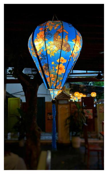 Photo of an illuminated Vietnamese lantern suspended from a dark ceiling in a hotel’s covered lobby. The lantern is shaped like a hot air balloon, with silk fabric stretched over bamboo ribs. It features a design of stylized orange-and-black koi fish and orange flowers on a vivid blue background. A long blue tassel hangs from the lantern bottom. Out of focus beyond the lantern are a slim tree trunk, tables and chairs, and a barrier made of doors of different sizes and colors joined together. Outside is a glimpse of orange lanterns hanging in the covered lobby of another hotel.