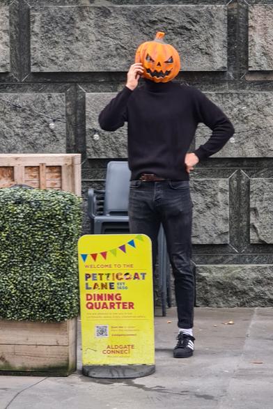 Someone wearing a mask in the shape of a Halloween carved pumpkin has a phone raised to their head, and is standing behind a sign saying "Welcome to the Petticoat Lane Dining Quarter"