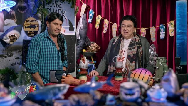 Päter und Olli in der neuen Folge "Zombiesharks". Päter mit schwarzer Perücke und Holzfällerhemd, Olli auch mit Perücke, einer grauen Jacke und einem Schal bekleidet.