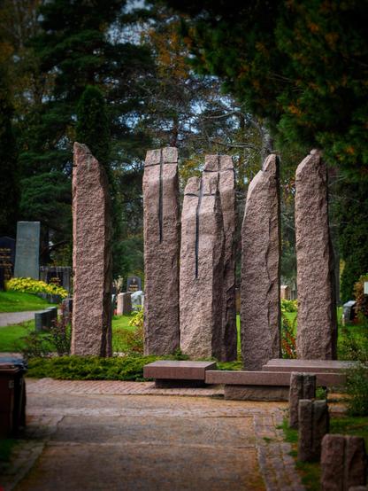 A cemetery scene featuring large granite monuments arranged vertically, with a pathway leading through them. Surrounding greenery and benches create a tranquil atmosphere.