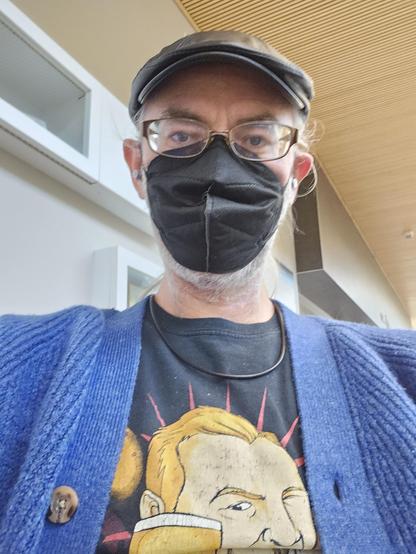 Waiting room selfie. I'm wearing my black leather flat cap and KN95, and a blue cardigan uver a Simon Pegg T-shirt (as Shaun, of Of The Dead fame). The ear loops on my mask pull my ears out sort of cocked, but I'm so used to it at this point I barely notice.