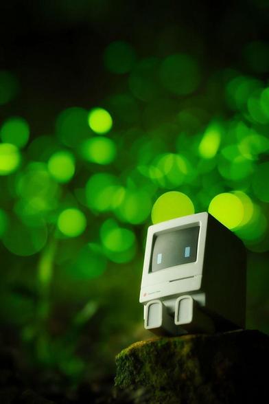 A small toy figure shaped like a retro computer with a screen displaying two square blue eyes sits on a mossy rock. The background is filled with soft, out-of-focus glowing green lights, creating a dreamy, whimsical atmosphere.