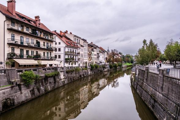 Blick über die Ljubiljanica, den Fluss, der durch Ljubljana führt. Links Häuser, rechts eine Promenade. Der Himmel ist bewölkt.