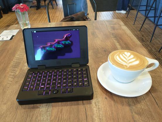 An MNT Pocket Reform mini laptop on a wood table with a latte and a glass containing a couple of fresh flowers.  A hardwood floor is visible behind.  The latte has tulip art with a heart-shaped flower head.