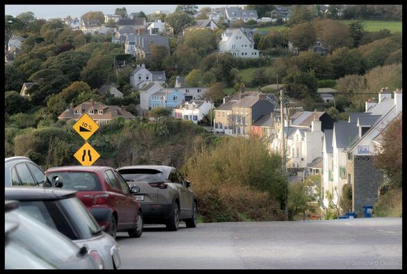 A steep residential street lined with parked cars descends towards a picturesque Irish coastal village with colourful houses nestled among mature trees on a hillside, with two yellow diamond-shaped road signs showing "10%" gradient warning and narrow road ahead symbol in the foreground