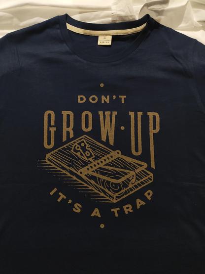 Blaues T-Shirt mit Print auf der Vorderseite.
Der Print besteht aus einer Mäusefalle, über welcher "DON'T GROW UP" und darunter "IT'S A TRAP" steht.