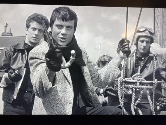 BW movie still of three white men standing up. The two in the back wear moto bike jackets and one has goggles on his helmet. In the middle Oliver Reed wearing a tweed coat and black gloves brandishes a knife with a cane handle as if it came from his umbrella. Reed’s hair is short tidy and he is young and lithe.