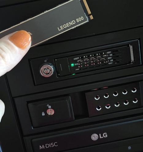 This is a close-up shot of a woman's hand holding an M.2 NVMe SSD in front of a black computer case.

* A woman's fingers, one with a sparkly orange-painted nail, are holding a black solid-state drive.
* The drive has "LEGEND 800" printed on it in white capital letters.
* Behind this, the front of a black computer tower or drive enclosure is visible.
* At the top is a hot-swappable NVMe drive bay with a key lock on the left and a green indicator light to the right.
* Below this bay is a hot-swappable SATA drive bay with a lock icon and a key lock on the left.
* At the very bottom, a portion of an LG optical drive is visible, with "M-DISC" branding.