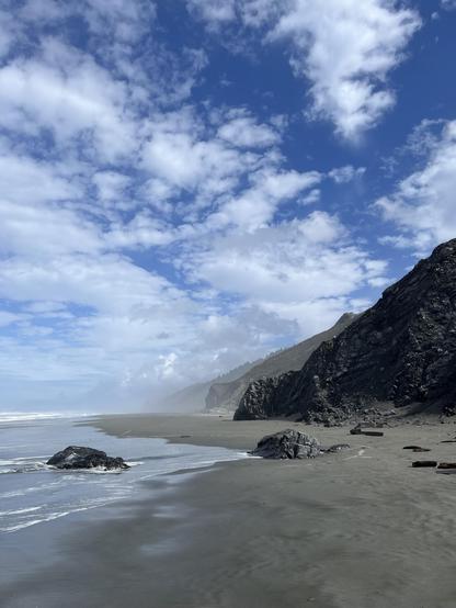 A mixture of clouds, fog, sun, and strong surf in the coastal section of Redwood National and State Parks. The park is open, though visitors may find a number of changes to available services during the shutdown. This image shows waves from the Pacific Ocean rolling onto the coast at about 41 degrees north latitude. The sand is warm from the sunlight and the sound of the waves is simply incredible. This image shows a series of rock outcrops along the beach (right side). In the foreground, a black and silver outcrop with veins of white. Behind this, an outcrop of green and grey color. In the mist in the distance, a large outcrop of green, black, and white rock forms a point of land that is generally impassable except during periods of very low tides. Harbor seals could be seen in the surf while overhead, migrating birds were already on the move.
