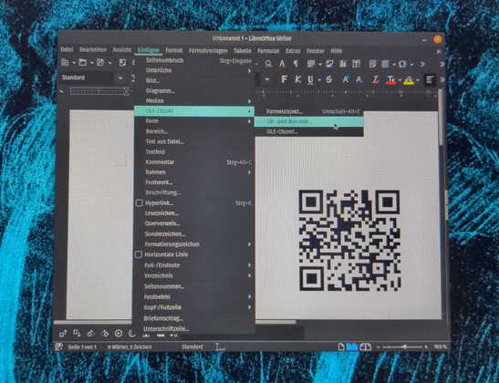 Ein QR-Code aus schwarzen Punkten und Quadraten auf weißem Grund im Fenster von LibreOffice Writer. Aufgeklappt das Programm-Menü, dass den Befehl zum Generieren und Einfügen eines QR-Codes enthält.