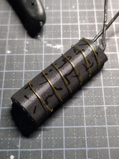 A dark gray plastic rectanglar pendent with black penmarks and golden wire wrapped around it in an artistic fashion with a black leather strapp emerging ffom it's top. It's laying on a dark gray knife mat with it's characteristic cross pattern. The habdle of some kind of tool is visible at the top of the picture.