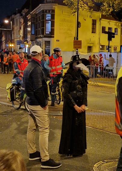 A female plague doctor dressed in black and with a raven face mask stands among the people from the emergency services, two on bikes. They are waiting in a lit street on the signal to walk on.