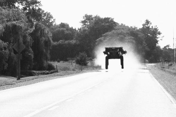 Looking down a road at an approaching farm machine that is trailing dust. The machine, a crop sprayer, is on large raised wheels.