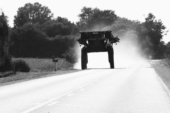 Looking at an approaching farm machine that is trailing dust. It is only a few car lengths ahead, approaching in the opposite traffic lane. The machine, a crop sprayer, is on large raised wheels.