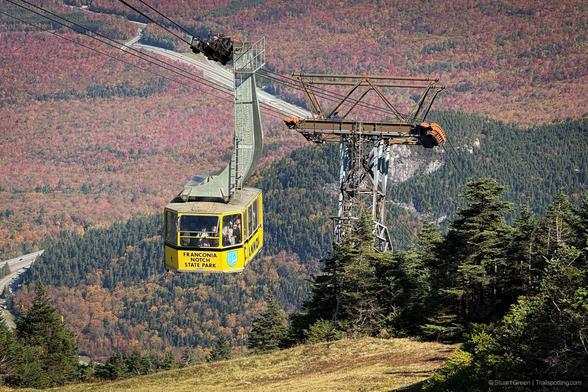 Yellow aerial tramway car labeled “Franconia Notch State Park” suspended on cables above a forested mountain landscape. Autumn foliage in shades of orange and gold blankets the valley below, with a winding road visible through the trees. Tramway tower and cables frame the scene, highlighting the park’s high-elevation transport system.