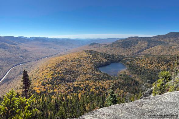 Panoramic autumn mountain landscape viewed from a high vantage point. A winding road traces the valley on the left, bordered by dense forest in shades of yellow, orange, and green. At center, a small heart-shaped lake nestles among the trees. Rugged mountain ridges stretch into the distance beneath a clear blue sky.