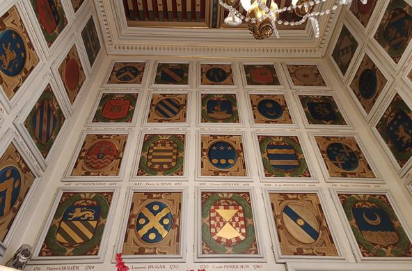 Photographie numérique couleur en format paysage, avec une vue en contreplongée d'un salon décoré sur toute la hauteur de ses murs de blasons enluminés. On est face à un des murs et on voit partiellement 2 autres murs.
On aperçoit également le plafond à la française richement décoré et un lustre doré à pamprilles (des guirlandes de verre).