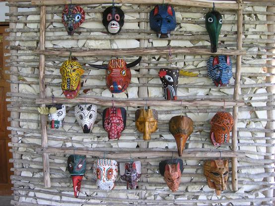 A white rock wall has long pieces of bamboo tied together to form a rack to hang masks from. There are 19 Mayan masks hanging, all different face designs and colors. Some of the masks represent animal faces, others seem to represent spirts. Several of them are brown, but they are all colorful. Guatemala