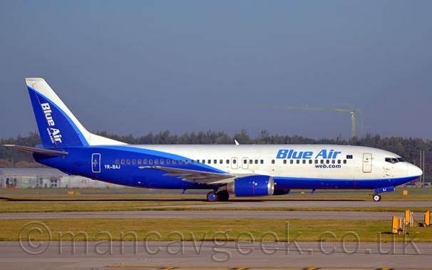 Side view of a twin engined jet airliner taxiing from left to right.
The plane is mostly white, with a blue belly at the front, although the rear is mostly blue with a white roof, with a light blue stripe separating the 2 sections.
There are blue "Blue Air" titles on the upper forward fuselage, above the passenger cabin windows, with smaller, darker "web.com" web address below the windows.
The white registration "YR-BAJ" is on the lower rear fuselage., just ahead of the aft cabin door.
The tail is mostly white, with a white leading edge, and white diagonal "Blue Air" titles and "Web.com" web address.
A grey runway and taxiway, lined with green grass, fills the foreground, while trees stretch across the background, under blue-grey sky.