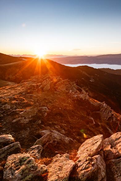 Das Bild zeigt eine beeindruckende Berglandschaft bei  Sonnenuntergang. Im Vordergrund sind große, kantige Felsen und eine felsige Wiese zu sehen, die vom warmen, goldenen Licht der Sonne intensiv angestrahlt werden. Die Sonnenstrahlen breiten sich fächerförmig über das Bild aus und tauchen die Szene in kräftige Orange- und Rottöne. Dahinter erstrecken sich dunkle Berge, deren Silhouetten gegen den hellen Horizont wirken. Weiter hinten, im Tal, liegt eine weiße Nebeldecke, die das Bild geheimnisvoll und atmosphärisch macht. Am linken Bildrand sieht man den sanften Abhang eines Berges, während am rechten Rand die Hänge steil nach unten abfallen. Ganz klein erkennt man auch eine Person, die auf den Felsen steht und in die Ferne blickt – das verstärkt die Dimensionen der Landschaft und den Eindruck von Weite. Das Bild vermittelt eine Stimmung von Ruhe, Erhabenheit und Naturgewalt, mit einem sehr starken Fokus auf das Lichtspiel der  untergehenden Sonne.