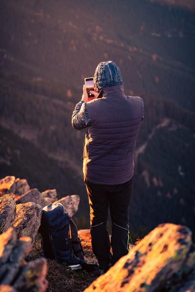 Auf diesem Bild sieht man eine Person, die auf einem felsigen Bergvorsprung steht. Sie trägt Outdoor-Kleidung: eine dunkle Hose, eine wattierte Weste sowie eine Kapuze wegen der Kälte am Abend. Die Person hält ein Smartphone in den Händen und fotografiert die Landschaft vor sich. Neben ihr am Boden steht ein großer Wanderrucksack, was darauf hindeutet, dass sie auf einer längeren Wanderung oder Bergtour unterwegs ist. Der Vordergrund ist von Felsen und Steinen geprägt, die im warmen Licht der tiefstehenden Sonne orange-golden leuchten. Im Hintergrund fällt der Blick steil in ein bewaldetes Tal hinunter. Die Bäume erscheinen dunkel und dämpfen den Kontrast zum hell angestrahlten Vordergrund.
Im vorherigen Bild ist die Person winzig klein auf den Felsen zu sehen. Hier ist sie im Detail dargestellt, mitten in diesem Moment des Innehaltens und Fotografierens.