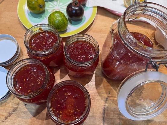 Large Mason jar plus four small jars with red tomato preserve on counter alongside lids and other plates with lemon and tomatoes.