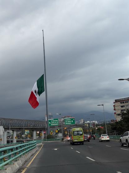 A view of a highway with a large Mexican flag at half-mast. There are traffic signs indicating directions, a green bus, and other vehicles on the road. The sky is overcast with dark clouds.