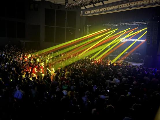 This photo captures an energetic live concert in a large indoor venue. The image shows a packed crowd of hundreds of attendees filling the floor space, with people standing densely together facing toward a stage area on the right side of the frame.

The lighting design is spectacular, featuring dramatic beams of colored light - primarily yellow, orange, and some red/pink tones - cutting diagonally across the venue from right to left. These sharp, parallel light beams create a dynamic visual effect through the atmospheric haze in the room, illuminating portions of the crowd in vibrant yellows and oranges while leaving other areas in darkness.

On the stage, there's visible branding or text that appears to read "DELTRON 3030." Professional lighting rigs and truss systems are visible mounted on the ceiling and walls.

The venue is the Majestic Theater in Detroit, a modern club with high ceilings, industrial-style architecture, and professional concert infrastructure. The crowd seems highly engaged, with some people visible with phones out capturing the moment.