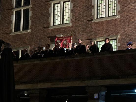 Het 3 October committee staat met het 3 October vaandel op het bordes van het stadhuis van Leiden tijdens het Reveille. Iedereen is in het zwart gekleed. Het is donker buiten. Uit de ramen van het stadhuis komt licht.